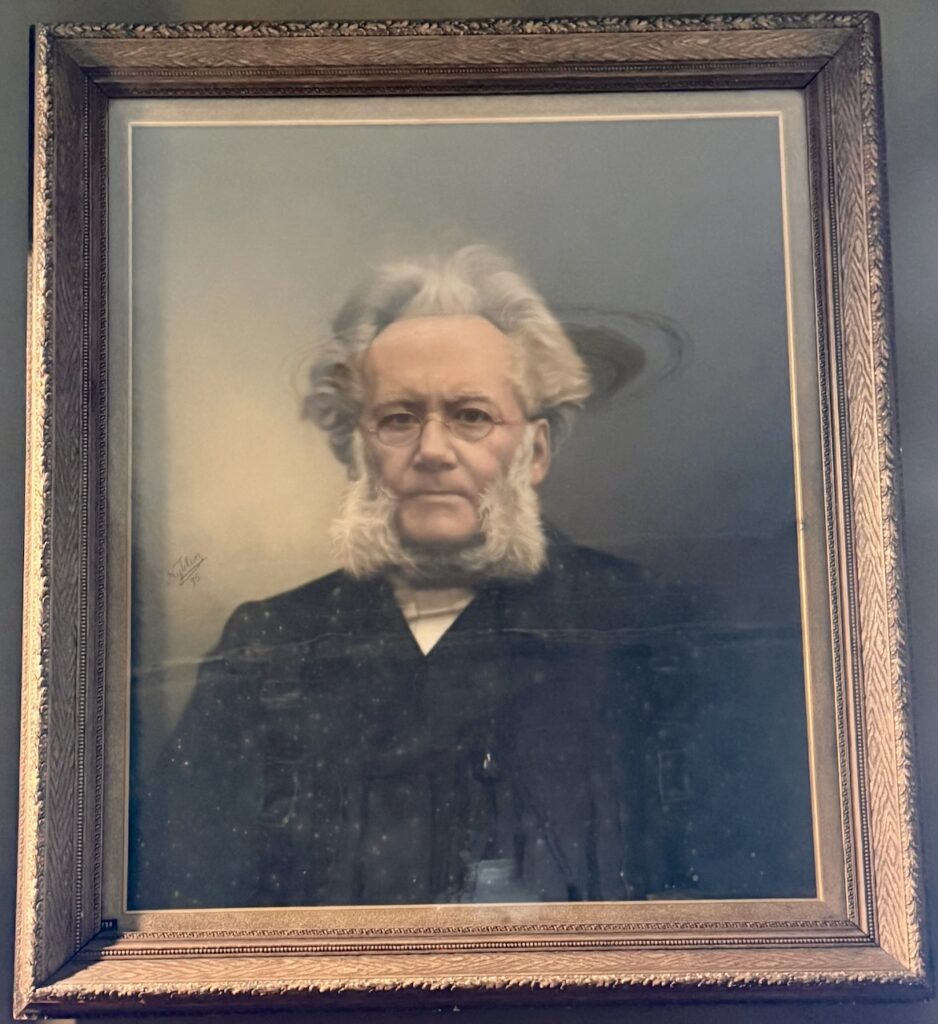 Wooden-framed portrait of Ibsen with prominent white hair, whiskers, and beard.