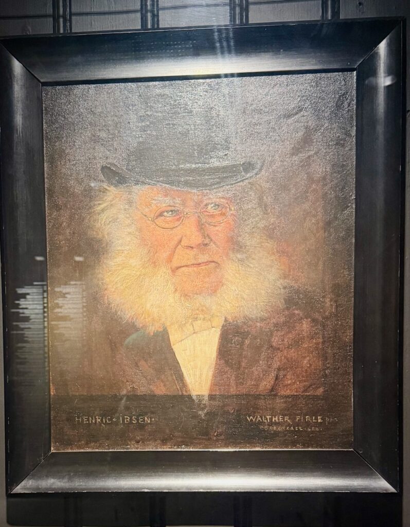 Portrait of Ibsen as an old man with white whiskers and beard, wearing glasses and a black top hat.