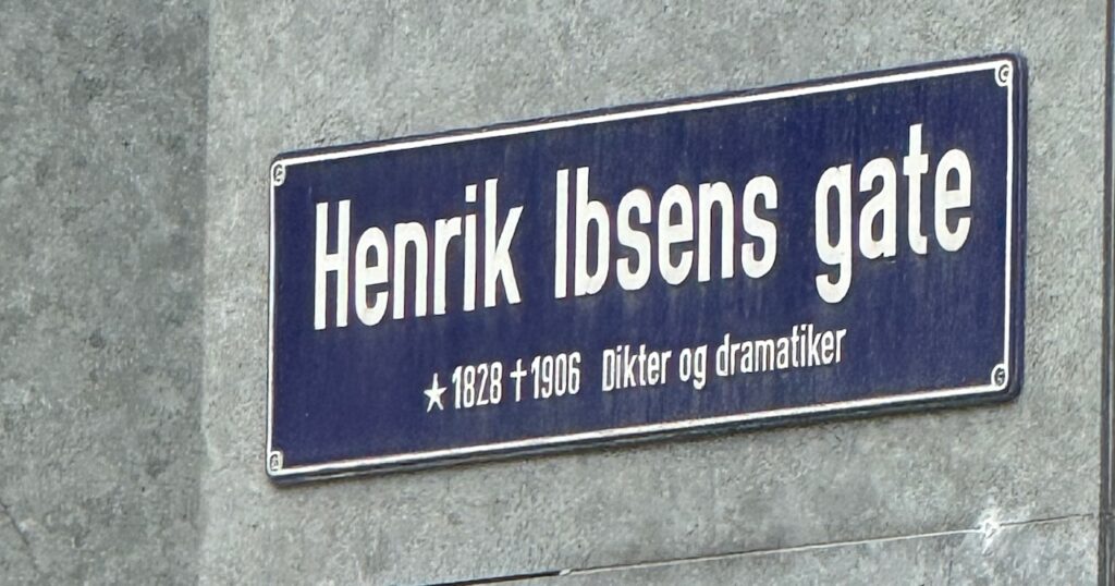 Street sign for Ibsen's Gate; dark blue sign with white lettering on the gray wall of a building.