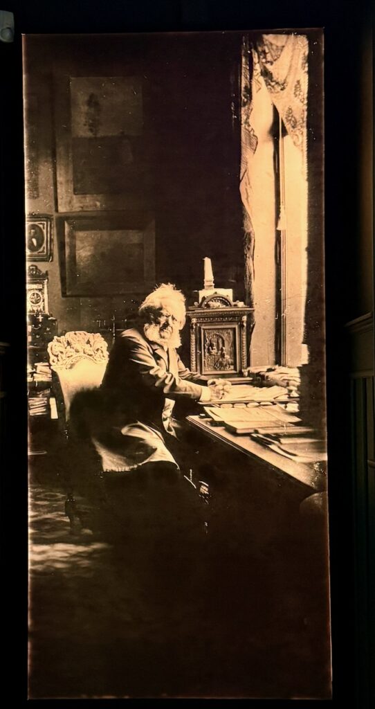 A photo of Henrik Ibsen sitting at his desk in his study. He is holding a pen and looking directly into the camera, as if distracted from the papers on the desk.