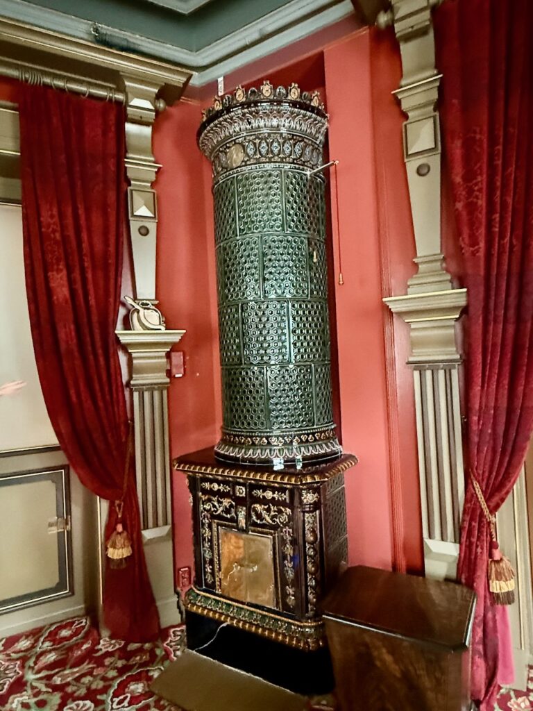 Tall ceramic stove in the corner of a room with red walls and red velvet curtains. The curtain frames are fancy gilt-painted columns.